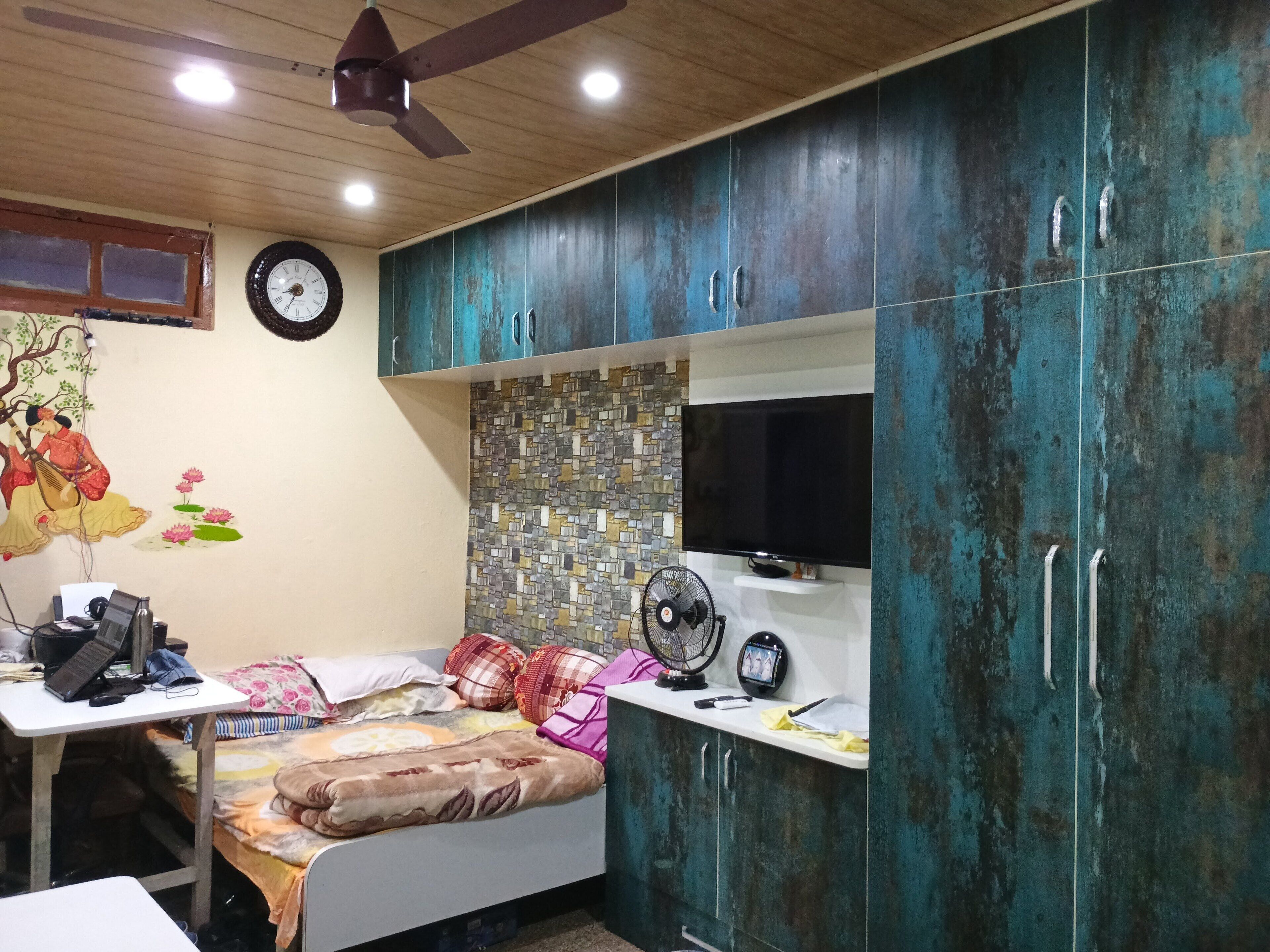 Un dormitorio con cama, escritorio, silla, ventilador de techo, televisor y armario.