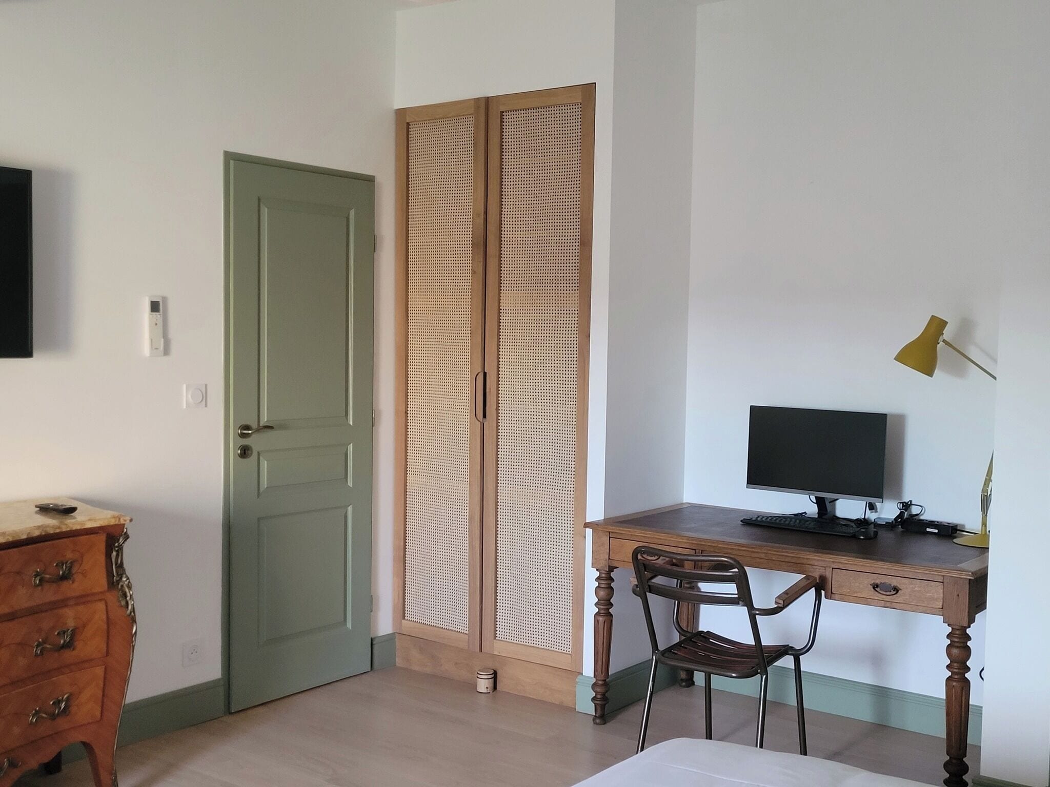 A room with a green door, a wooden desk with a computer, a chair, and a dresser.