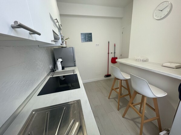 A modern kitchen with a built-in oven, a sink, and a countertop with a kettle and a coffee maker. There are two white bar stools and a red broom leaning against the wall. - Airtegal Black Sands (Cedeira)