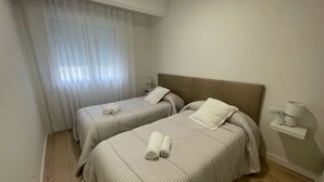 A hotel room with two beds, each with a checkered bedspread and rolled towels. There is a window with sheer curtains, a bedside table with a lamp, and a small shelf with a lamp and decorative items. - Airtegal Black Sands (Cedeira)