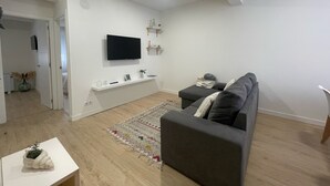 A modern living room with a grey sofa, a flat-screen TV mounted on the wall, a small white table with a plant, and a patterned rug. - Airtegal Black Sands (Cedeira)