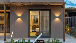 Een modern gebouw met een glazen deuropening, twee wandlampen en een terras met een lounge stoel en planten.