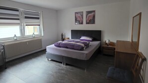 Een slaapkamer met een bed, nachtkastje, stoel, radiator en raam met zonwering.