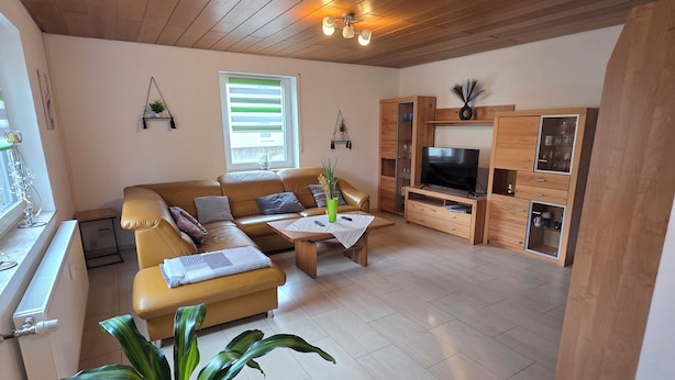 Een ruime woonkamer met een houten plafond, een gele bank, een salontafel en een tv-meubel.