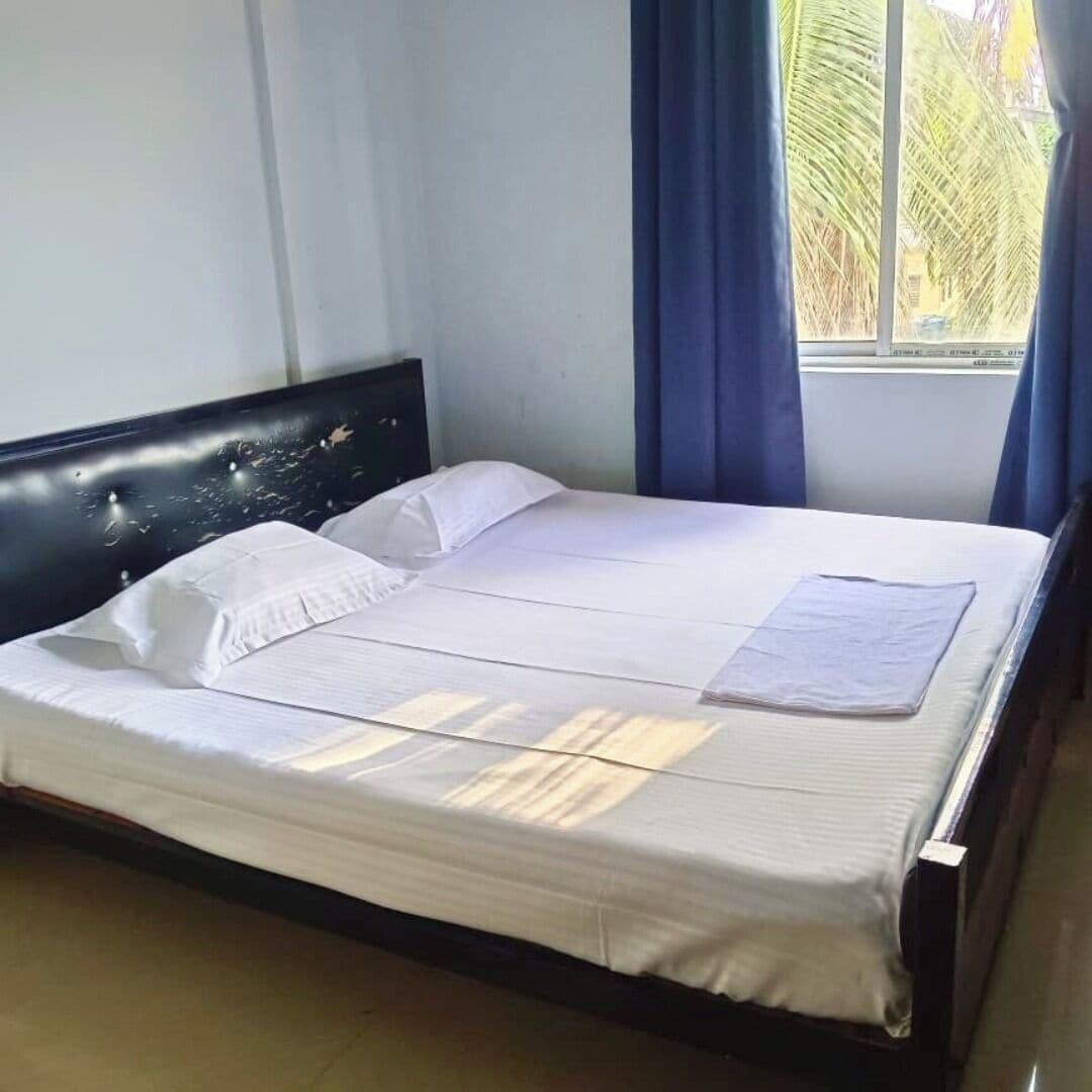 Un lit bien fait, avec du linge de lit blanc et une tête de lit, une fenêtre avec des rideaux et une vue sur des palmiers à l’extérieur.