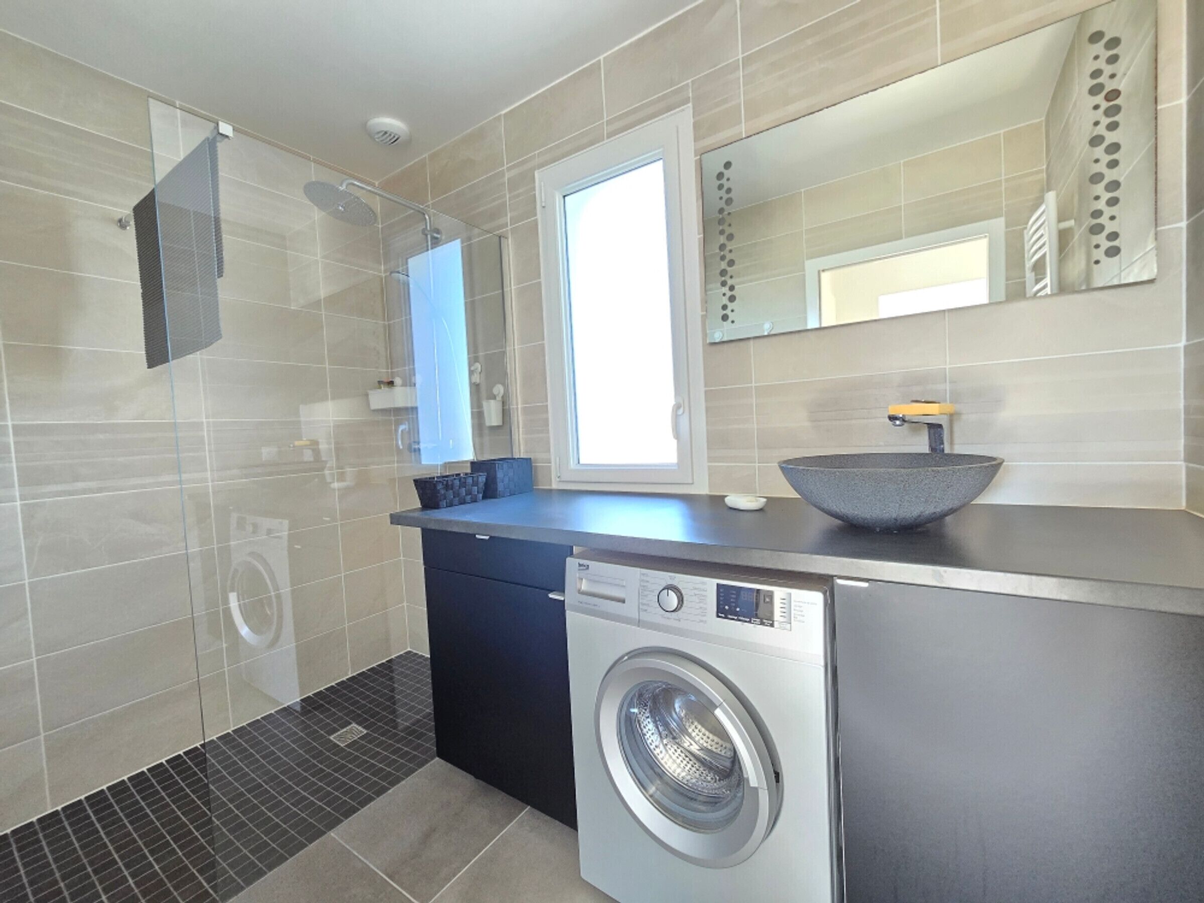 A modern bathroom with a built-in washing machine, a glass shower enclosure, and a large mirror above the sink.