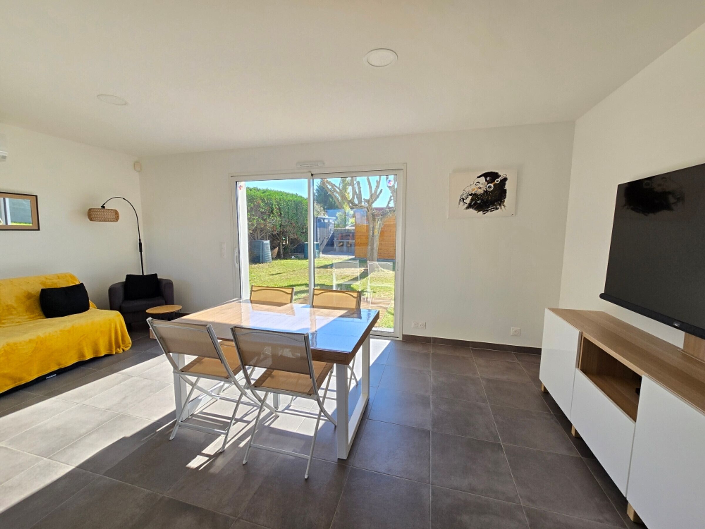 Un salon moderne avec un canapé jaune, une table à manger avec des chaises, un téléviseur et une porte-fenêtre coulissante donnant sur un jardin.