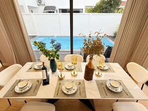 Een eettafel gedekt voor vier personen, met witte borden, gouden bestek en een fles wijn. Op tafel staat een vaas met gedroogde bloemen.