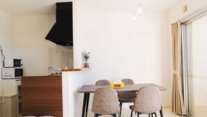 A modern kitchen with a microwave, sink, and black hood. There is a dining area with a table and chairs, and a window with curtains. - LoveOkinawa-Nakadomari (Onna)
