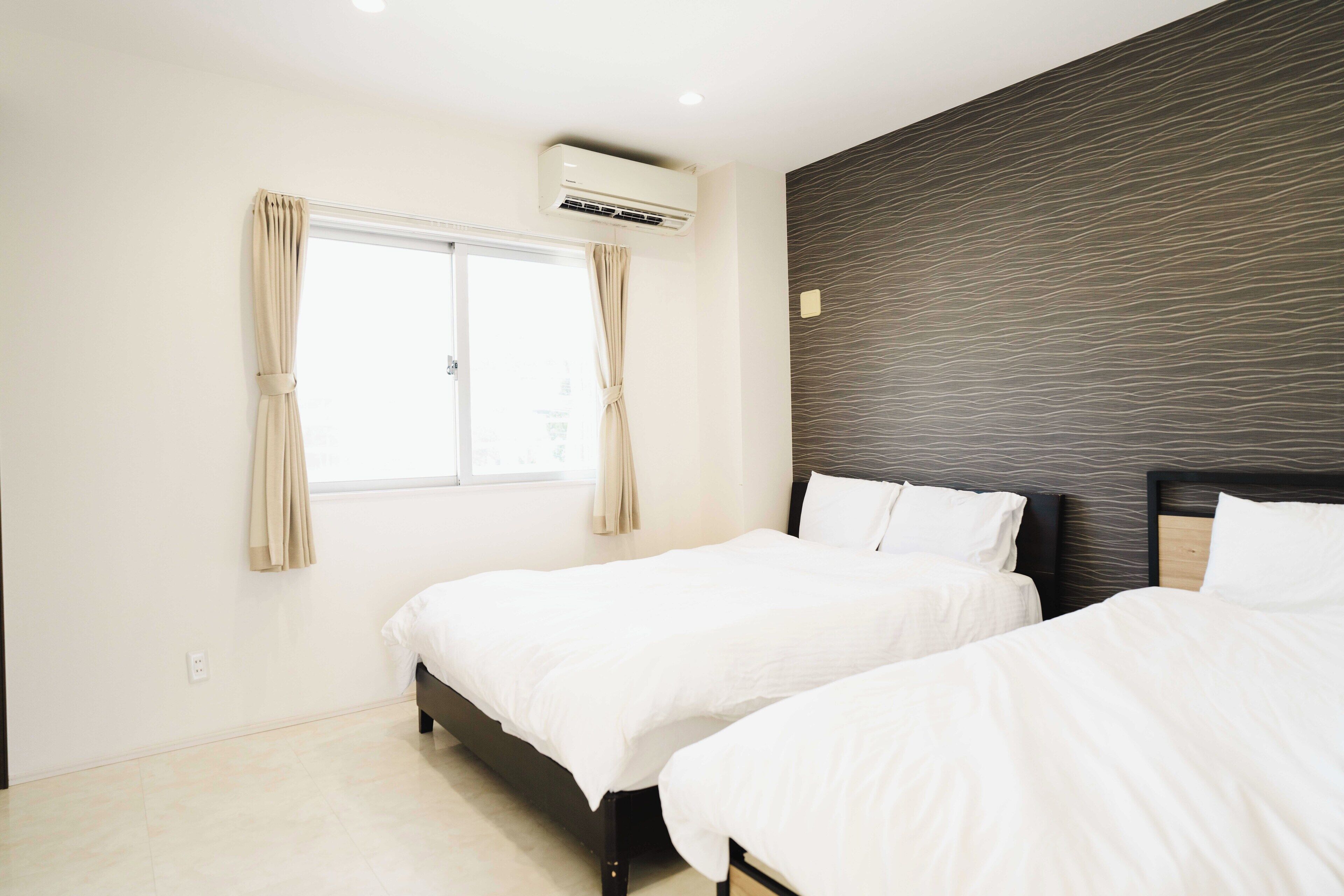 Une chambre d’hôtel avec deux lits simples, une fenêtre avec des rideaux, un appareil de climatisation et un mur à texture.