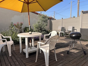 A patio with a round table, white chairs, a yellow umbrella, and a barbecue grill.