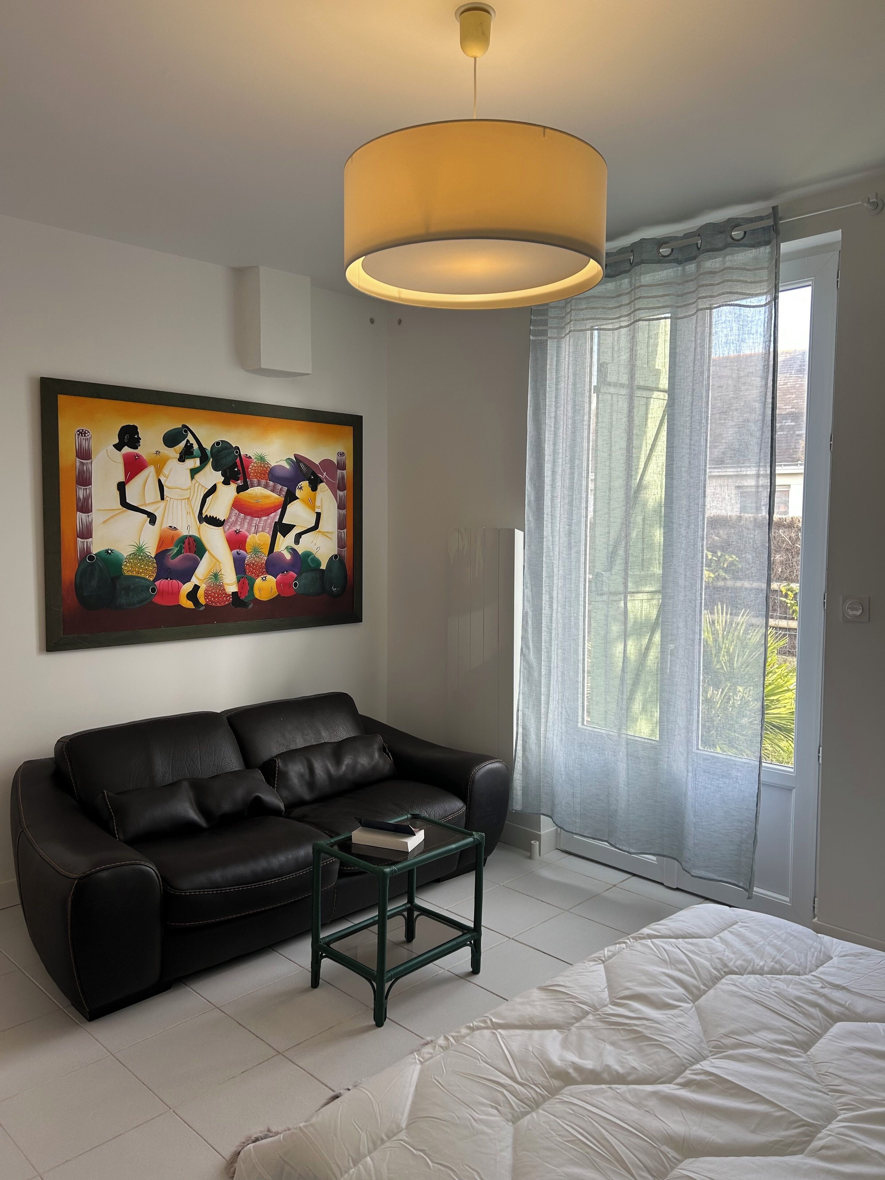 Une chambre moderne avec un canapé en cuir noir, un lit blanc, une petite table, un grand tableau et une fenêtre avec des rideaux.