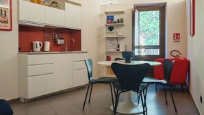 Een kleine keuken met witte kastjes, een rode aanrecht en een houten plafond. Er is een eethoek met een ronde tafel en stoelen, en een raam met uitzicht op groen.