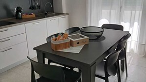 A modern kitchen with white cabinets, a black countertop, and a dining table with chairs. There is a clock on the wall and a window with sheer curtains.