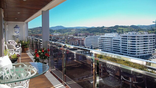 A balcony with a glass railing, white outdoor furniture, and potted plants.