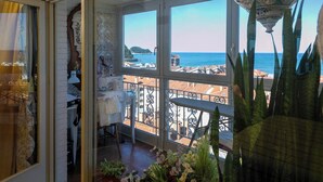 A balcony with a view of the sea and buildings, a table with a white tablecloth, and a potted plant.