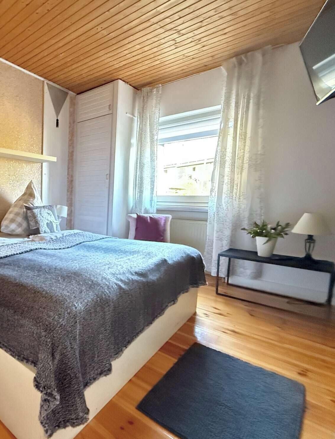 A bedroom with a bed, a nightstand, a lamp, a window with curtains, and a wooden ceiling.