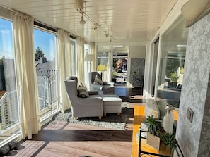 Een moderne woonkamer met een bank, een salontafel en een lamp. Er is een groot raam met gordijnen, een balkon en uitzicht op de omgeving.