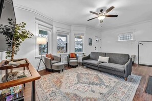 A living room with a gray sofa, two armchairs, a coffee table, a floor lamp, a ceiling fan, and a large window with a view of houses.