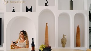 A woman sitting at a round wooden table with a wine bottle and a bowl of grapes. The room has white walls with arched niches containing decorative vases. - Sicilian Luxury for Families, 3 Minutes from the Sea and the UNESCO Cathedral. (Cefalù)
