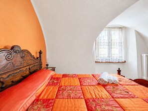 A bedroom with a wooden headboard, a quilted bedspread, a window with lace curtains, and a rolled towel on the bed.