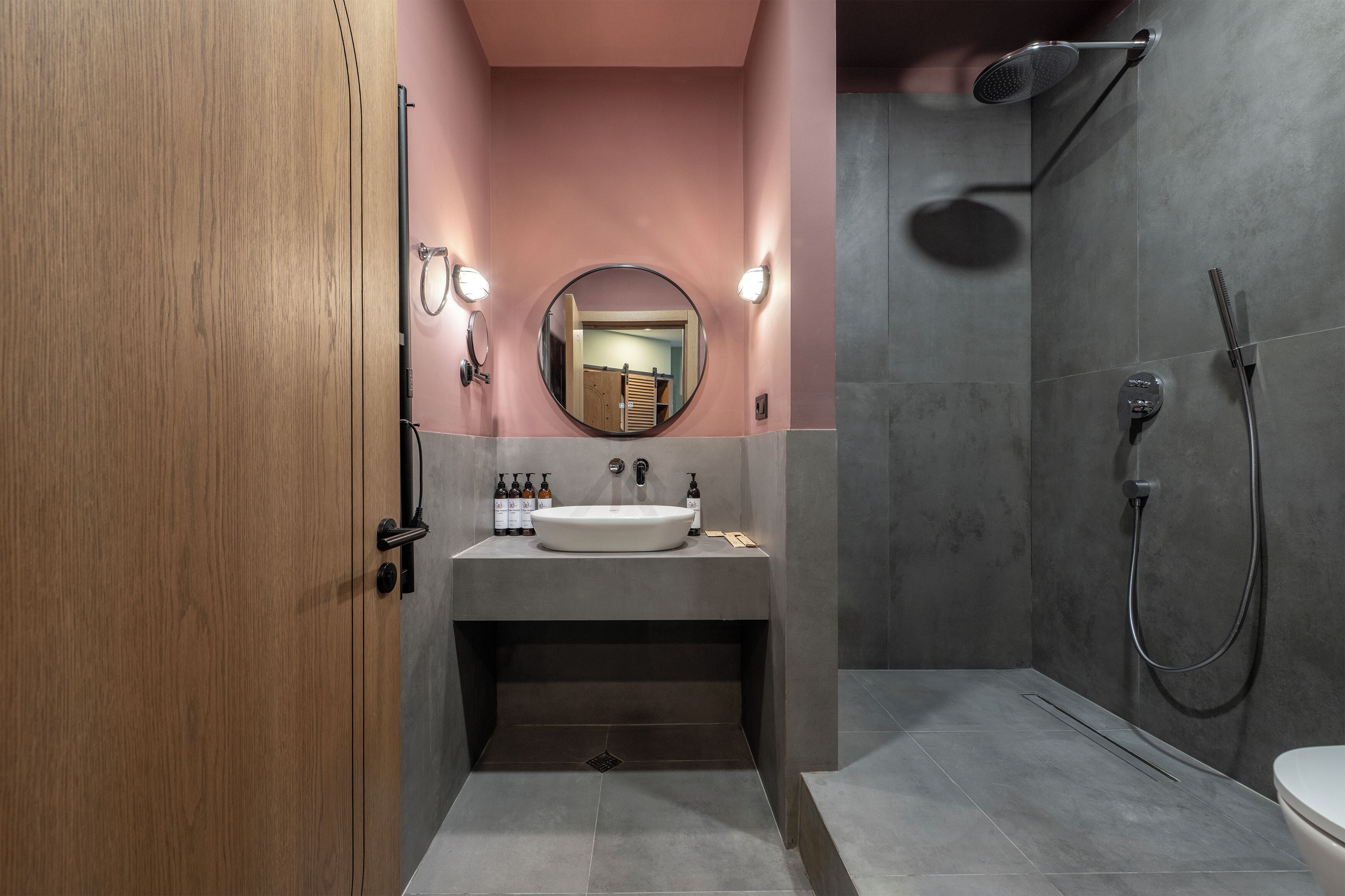 Un bany modern amb una porta de fusta, un mirall rodó, un lavabo i una dutxa.