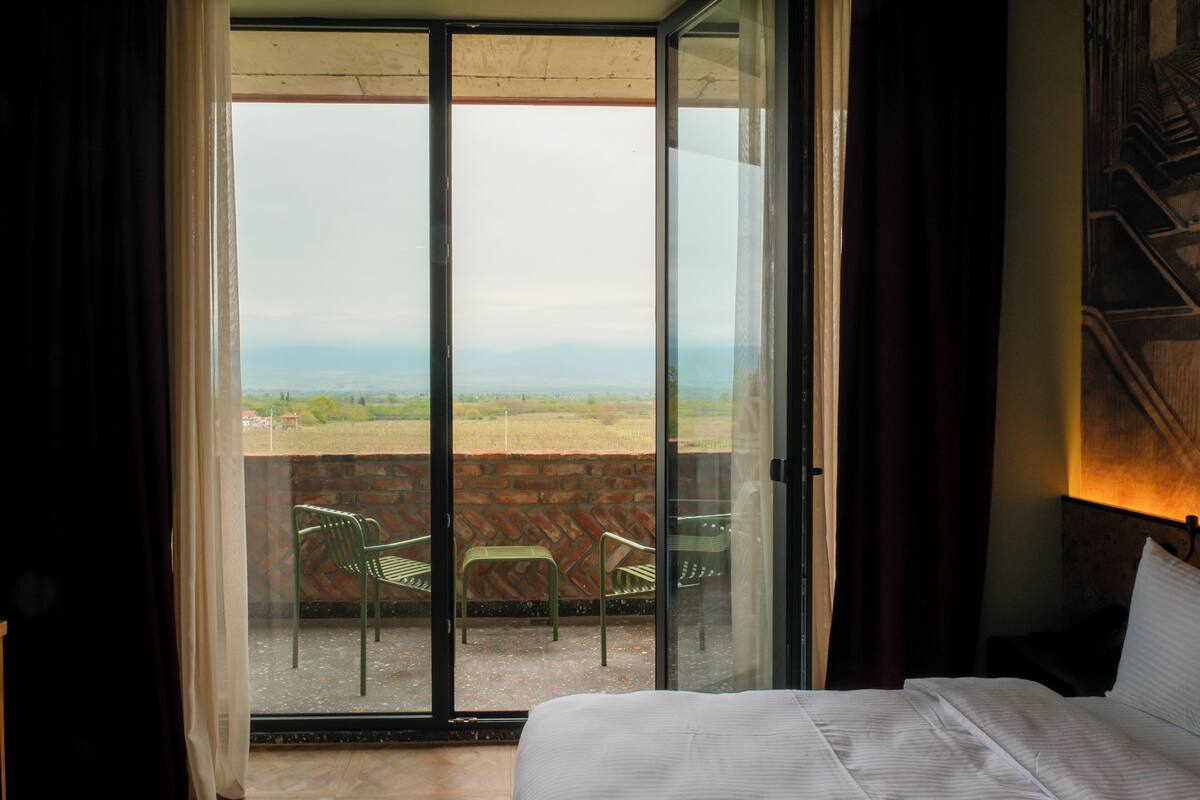 A room with a bed, a view through a sliding glass door to an outdoor area with chairs, and a brick wall.