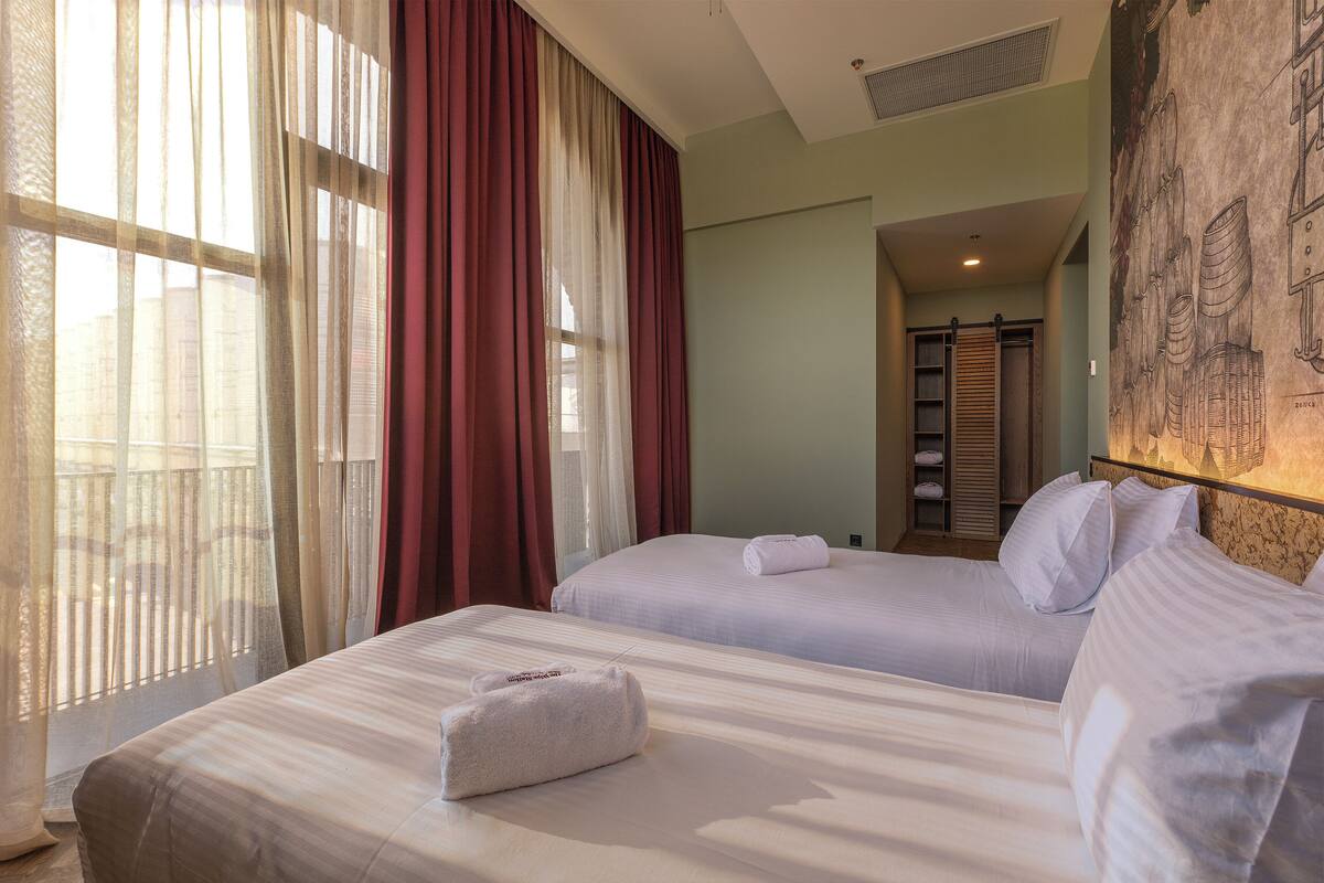 A hotel room with two beds, a large window with sheer curtains, a painting on the wall, and a hallway with a wooden door.