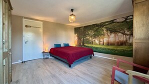 Ein Schlafzimmer mit einem Bett, einer Tür, einer Wandlampe und einem großen Fenster mit einer Waldmalerei.