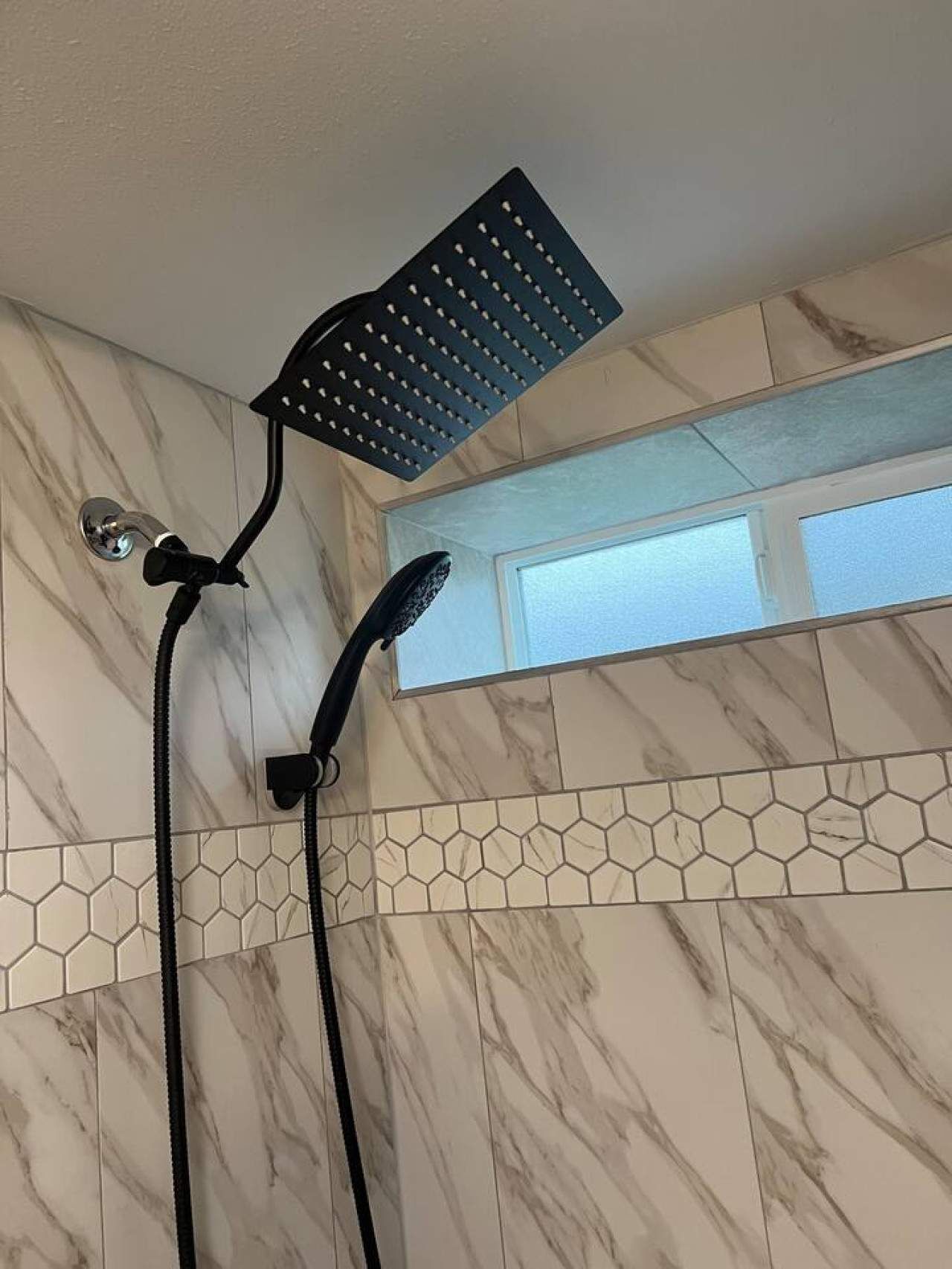 Une douche équipée d’un pommeau de douche à effet pluie et d’un pommeau de douche amovible, le tout installé sur un mur recouvert de carreaux hexagonaux et arborant un motif évoquant le marbre.