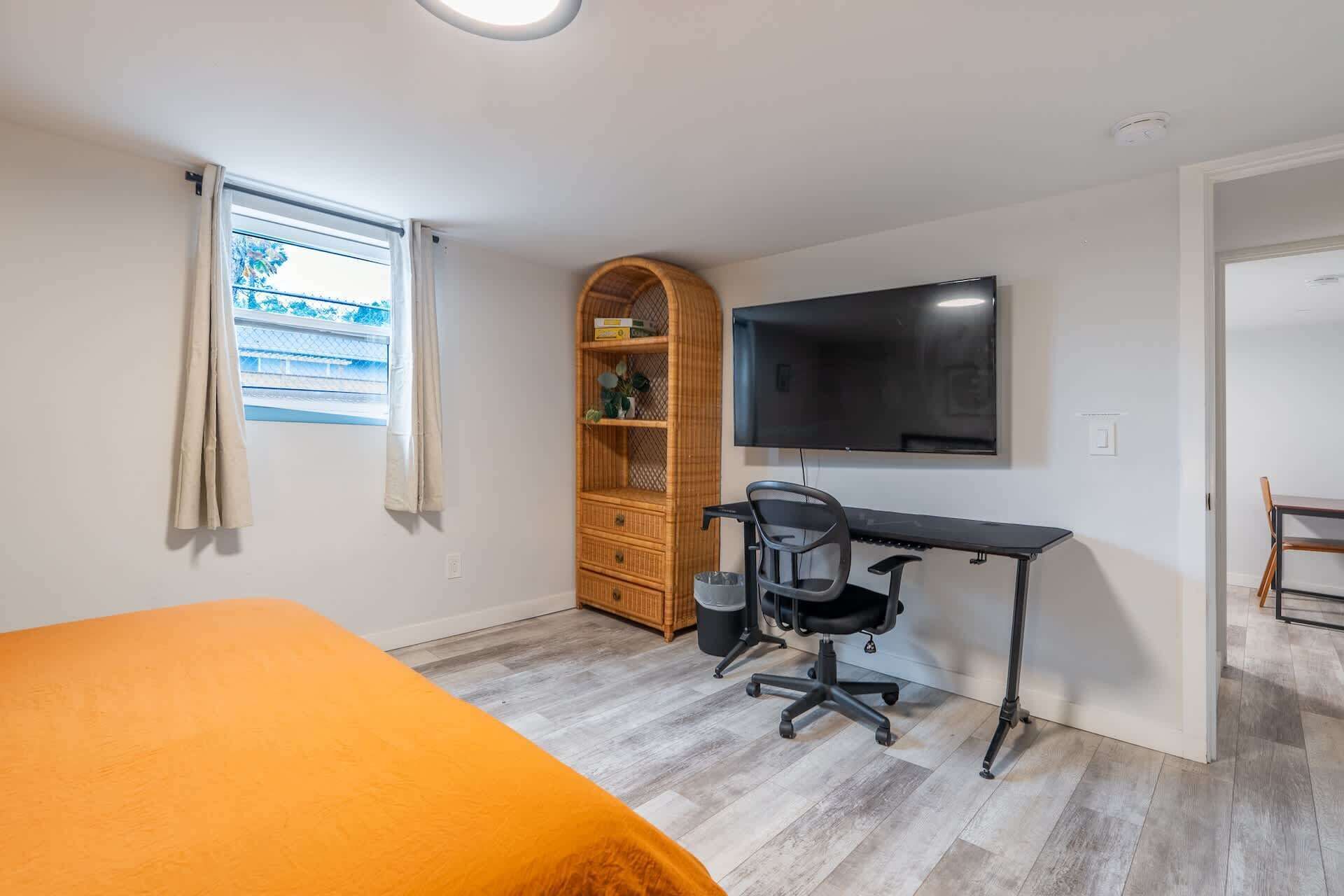 A bedroom with a bed, a desk with a chair, a TV, and a window with curtains.