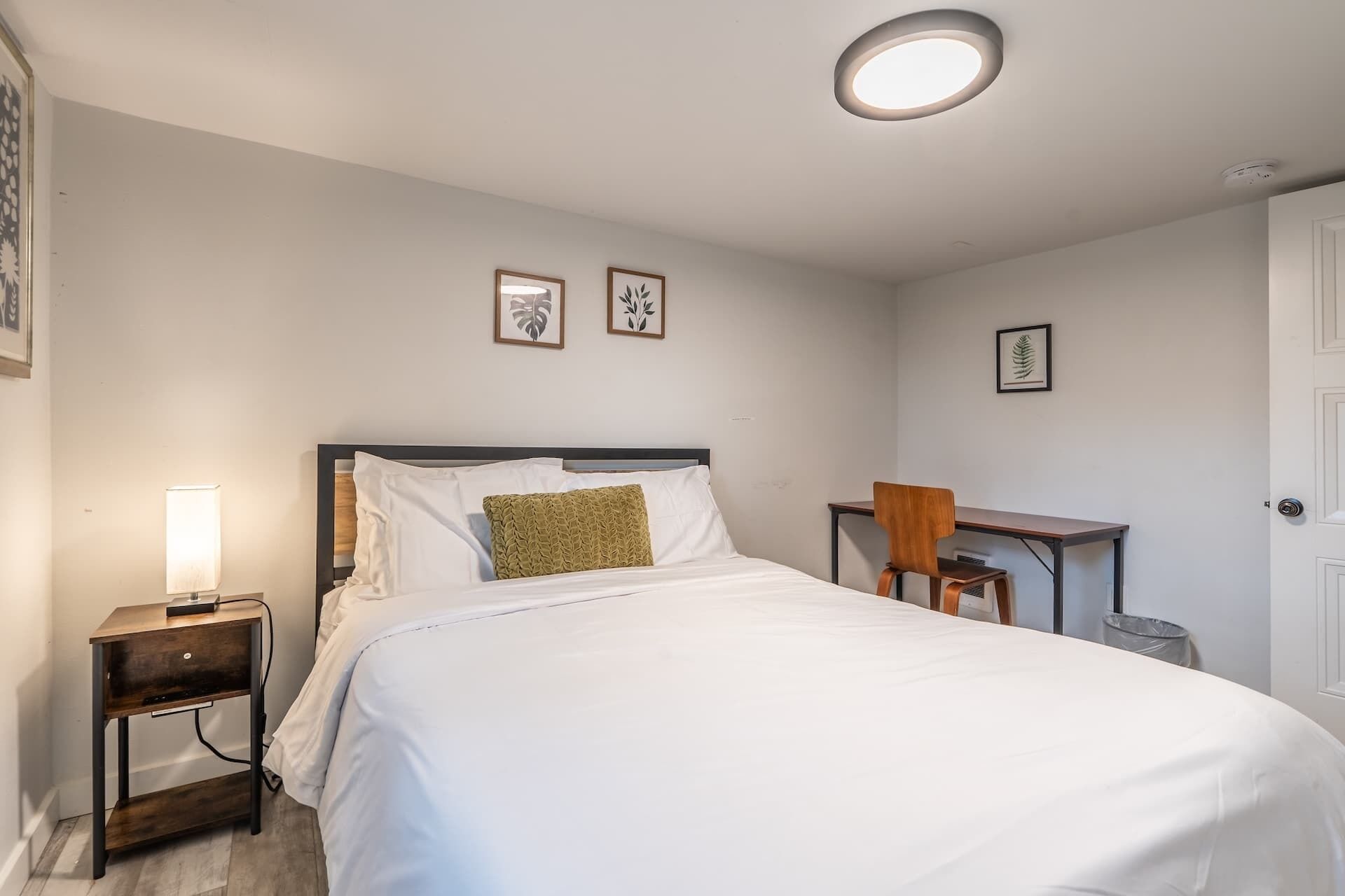 A neatly arranged bedroom with a bed, bedside table, chair, desk, and framed pictures on the wall.