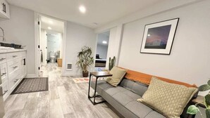 Una sala de estar moderna con un sofá gris, una mesita y una planta en maceta. Un pasillo da acceso a un baño.