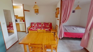 A compact room with a wooden table and chairs, a red and white checkered sofa, a single bed with a pink bedspread, and a wooden shelf with books.