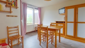 A cozy room with a wooden table and chairs, a bookshelf, a TV mounted on the wall, and a large window with curtains.