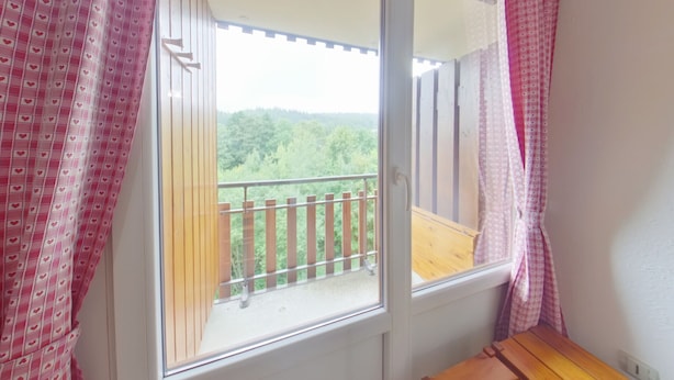 A room with a balcony, a wooden bench, and a view of a forested area.