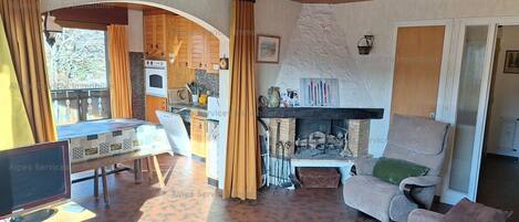 Un salon chaleureux avec une cheminée, un canapé, un téléviseur et un coin cuisine visible à travers une arche.