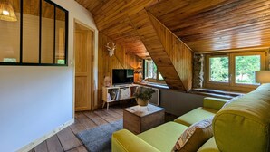 A cozy attic room with wooden beams, a television, a sofa, and a window with a view of greenery. - Renovated cottage for 4 pers. with equine accommodation, Saint-Front (Saint-Front)