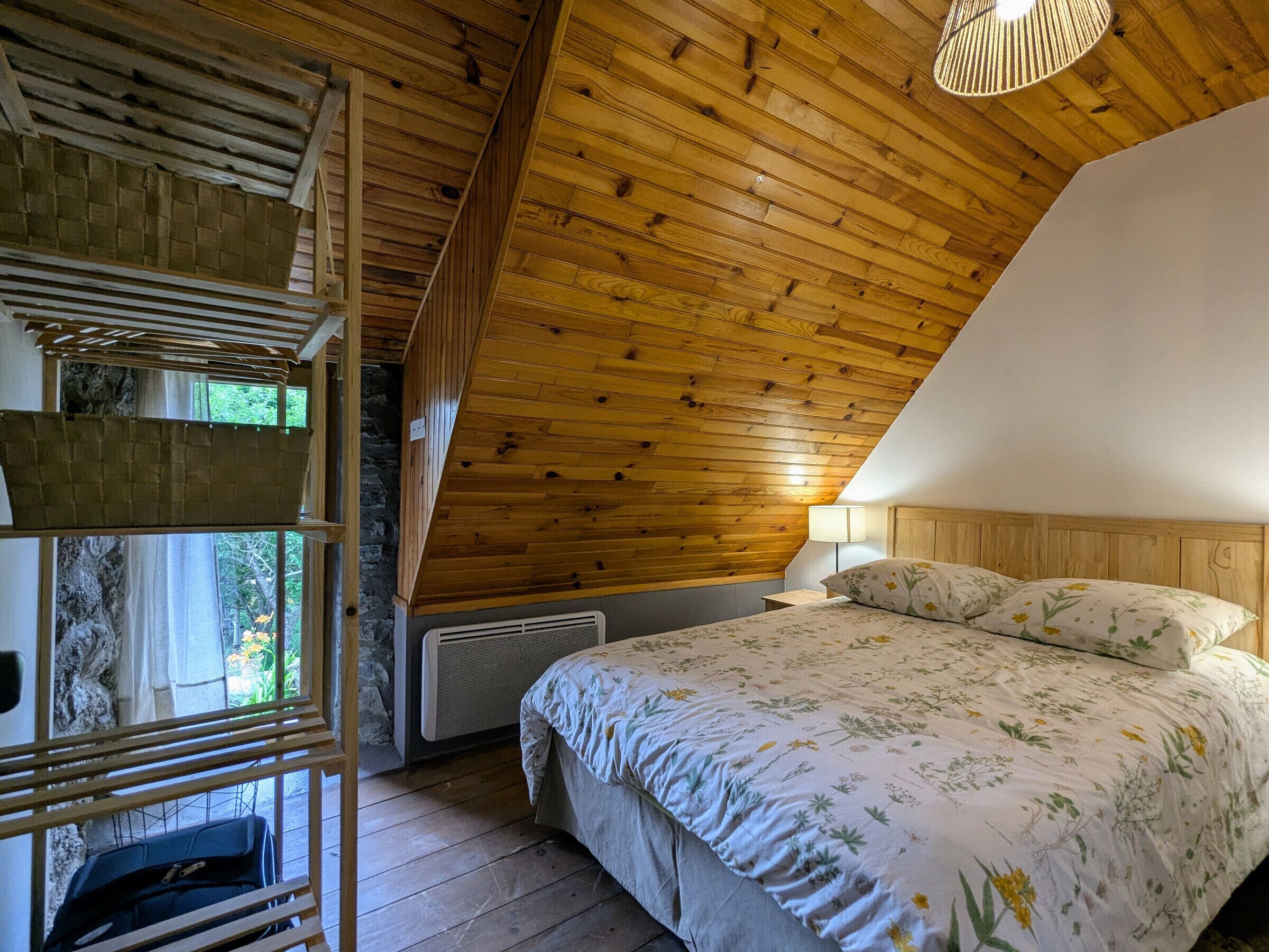 Un dormitorio con techo inclinado, vigas de madera, cama con ropa de cama floral, una lámpara de noche y una ventana con vistas a follaje.