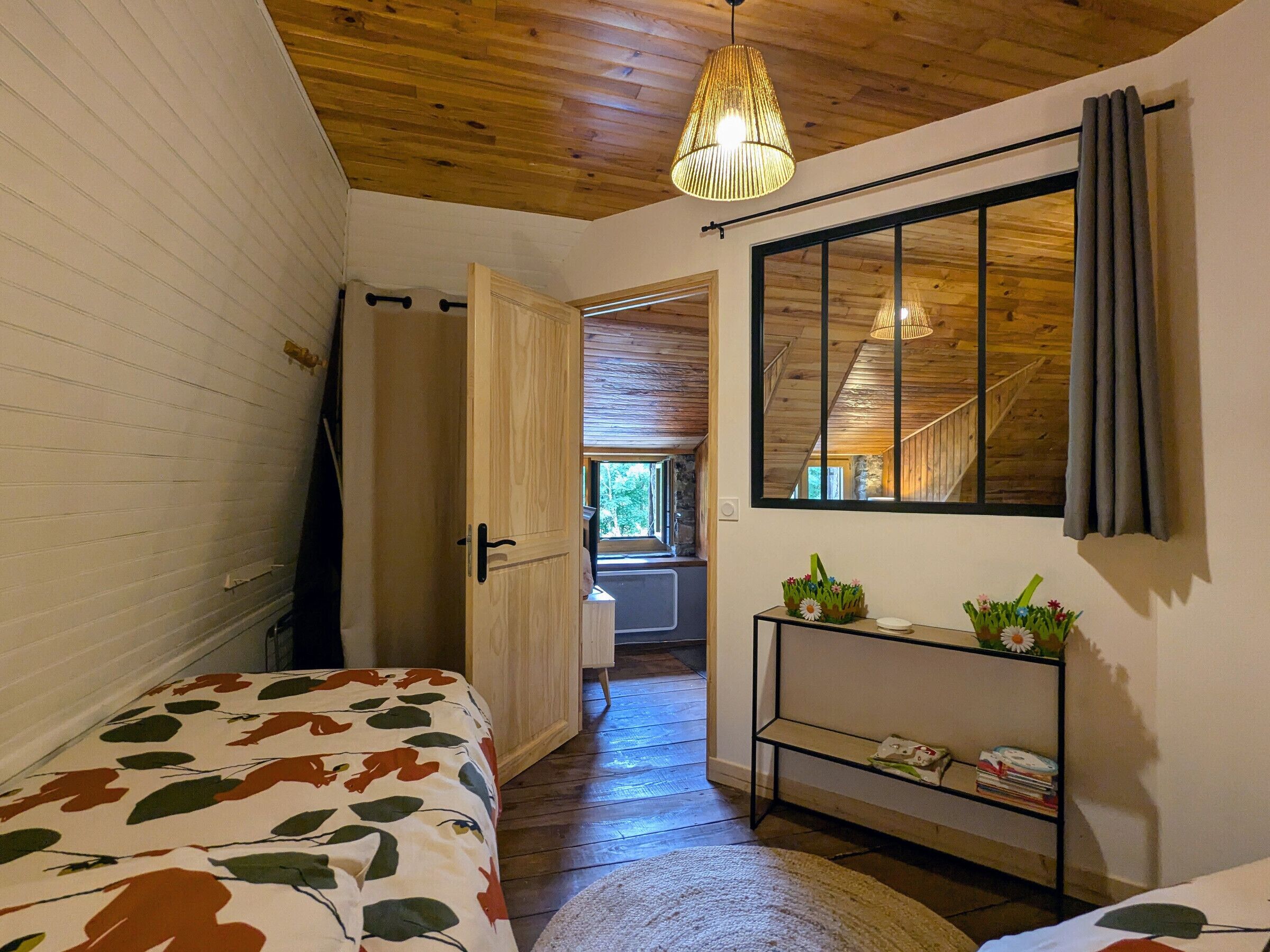 A bedroom with a wooden ceiling, a bed with a leaf-patterned bedspread, a bedside table with a mirror, and a shelf with books and flowers.