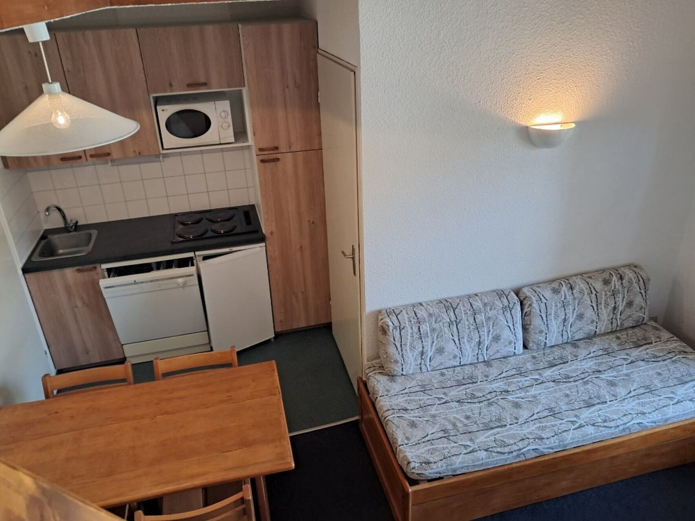 Eine kompakte Küchenzeile mit Herd, Mikrowelle und Spüle, ein Holztisch und eine Couch mit gemustertem Bezug.