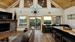 A spacious living area with a high ceiling, wooden beams, a stone fireplace, a large TV, a cozy seating area, and a dining table with chairs.