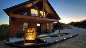 A wooden cabin with a porch, a sign reading "COZY HOLLOW," and a gravel driveway.