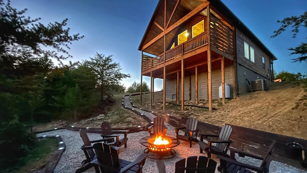 A two-story wooden cabin with a deck, surrounded by trees and a gravel path leading to a fire pit with chairs.
