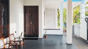 A covered entrance with a dark wooden door, a seating area with wooden chairs, and a potted plant.