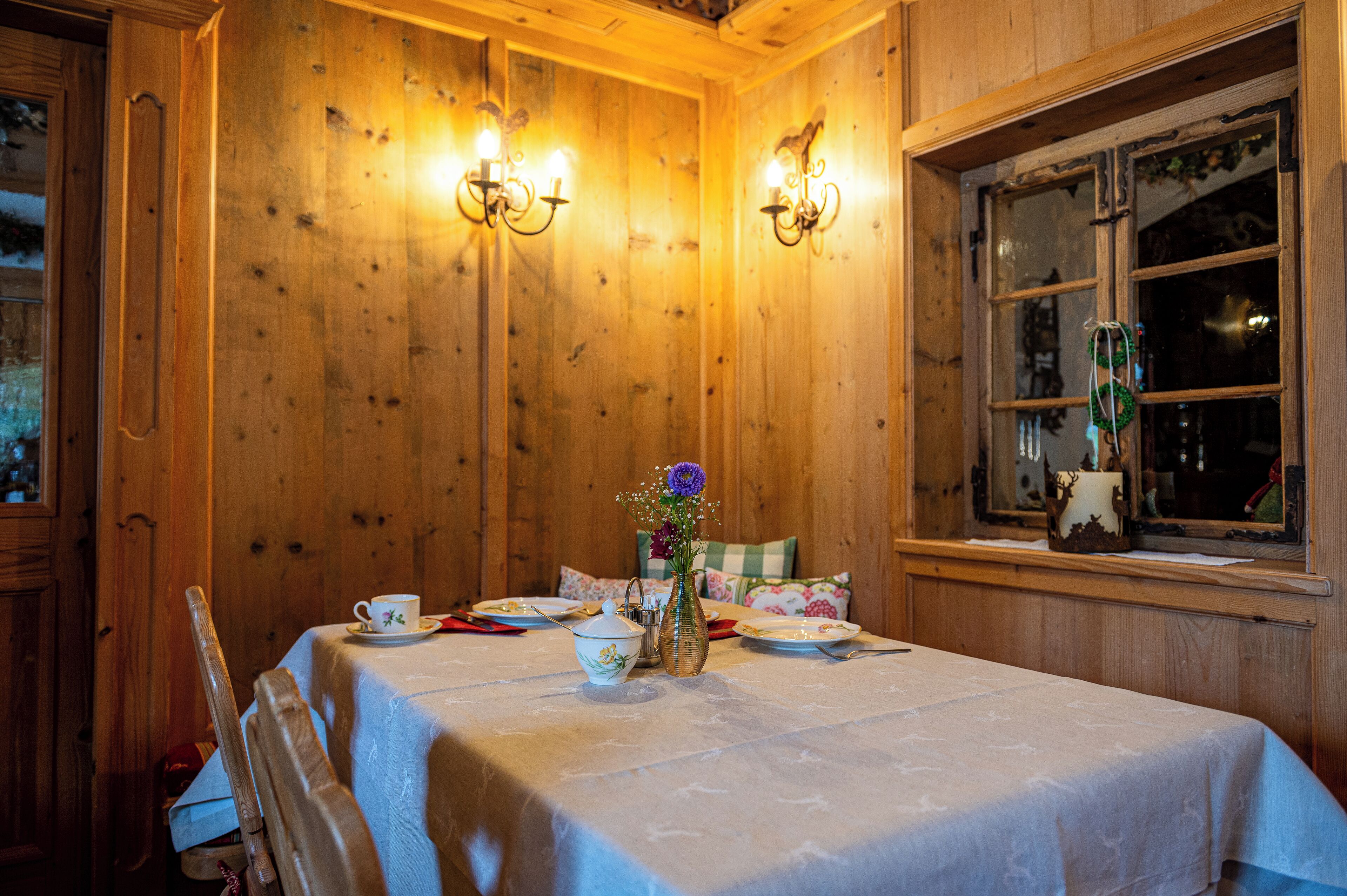 Um quarto com painéis de madeira, uma mesa de jantar posta para dois, um vaso com flores e uma janela com espelho e enfeites.