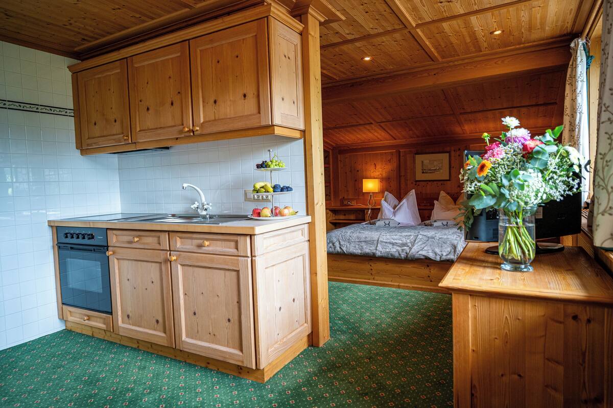 A wooden cabin interior with a kitchen area featuring a dishwasher, wooden cabinets, and a sink. There is a bed with a green patterned bedspread and a vase with flowers on a wooden table.