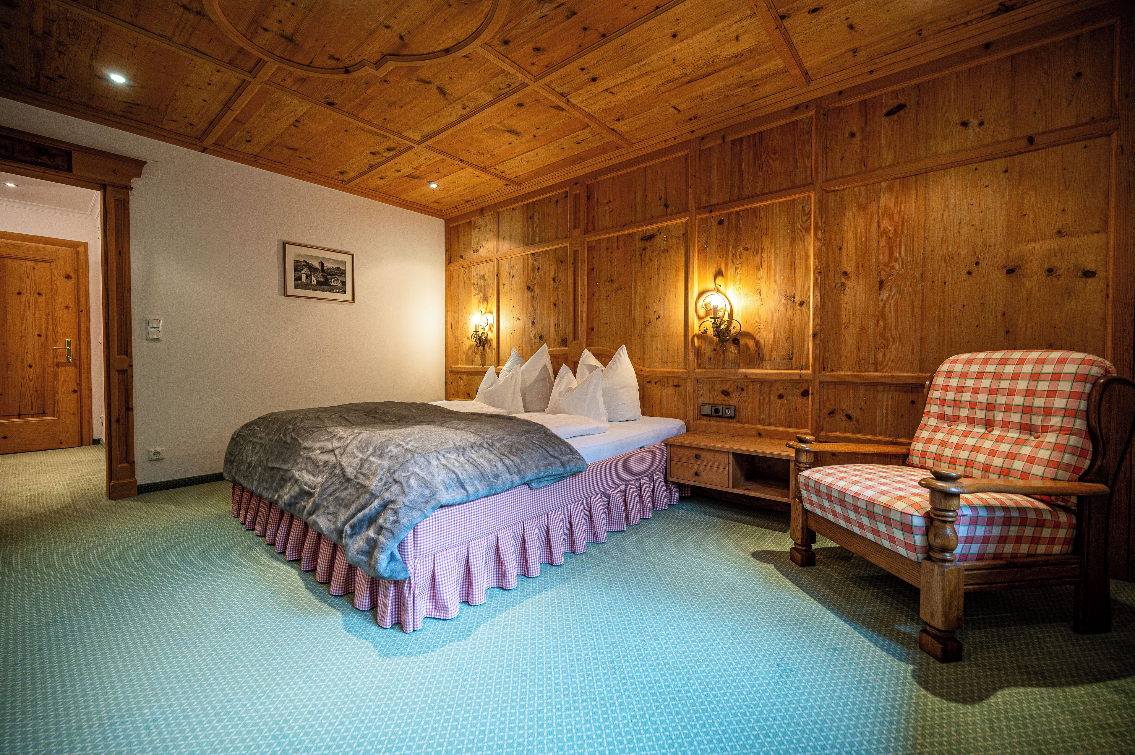 A bedroom with a wooden ceiling, a bed with a checkered bed skirt, a wooden chair, and wall-mounted lights.