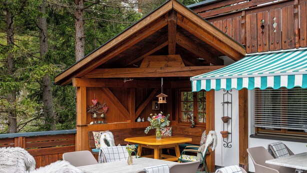Una cabaña de madera con zona de comedor que incluye una mesa y sillas, rodeada por una terraza de madera y árboles.