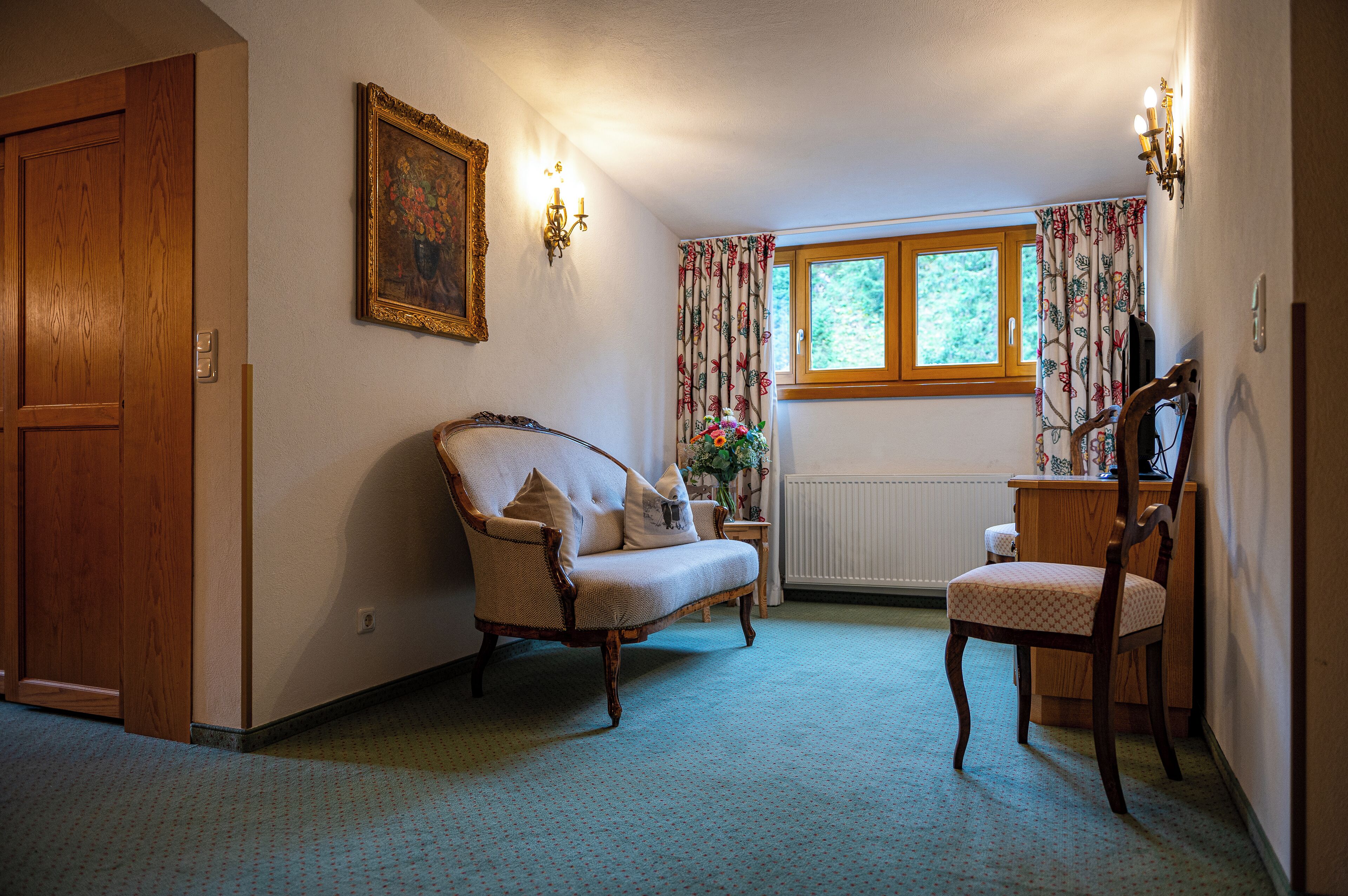 A room with a wooden door, a cushioned armchair, a wooden chair, a small table, and a window with floral curtains.